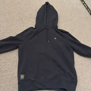Mörkblå hoodie från Morris - Säljer en stilren mörkblå hoodie från Morris. Den har en liten broderad detalj på bröstet och en justerbar huva med dragsko. Perfekt för en avslappnad look. Säljer på grund av att den är för liten.