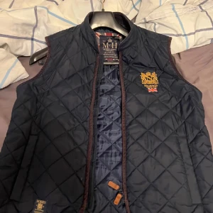 Mörkblå quiltad väst från Morris - Snygg mörkblå quiltad väst från Morris med broderad logga och brittisk flagga på bröstet. Västen har dragkedja framtill och en stilren design som passar perfekt för höst och vår. Perfekt för en casual look!