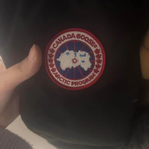 Svart dunjacka från Canada Goose - Säljer en svart dunjacka från Canada Goose med klassisk logga på ärmen. Jackan har dragkedja och knappar framtill, perfekt för kalla dagar. Den är designad för att hålla dig varm och bekväm under vintern.