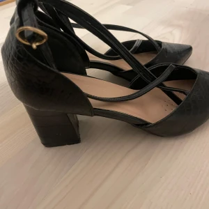 Svarta klackskor med remmar - Eleganta svarta klackskor med krokodilmönster och remmar. Skorna har en blockklack och en spetsig tå, vilket ger en stilren look. Perfekta för en kväll ute eller en speciell tillställning.