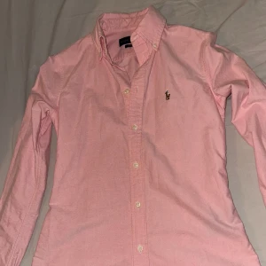 Rosa skjorta från Ralph Lauren - Snygg rosa skjorta från Ralph Lauren i storlek XS. Skjortan har en klassisk krage och knappar framtill. Perfekt för en stilren look.