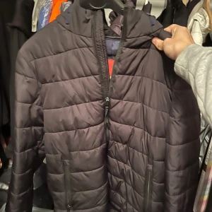 Svart quiltad jacka från Springfield - Säljer en svart quiltad jacka från Springfield i storlek M. Den har en dragkedja framtill och är perfekt för höst och vinter. Jackan är varm och har en stilren design med huva. Perfekt för kyliga dagar!
