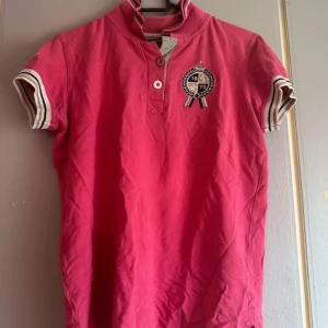 Rosa pikétröja från Copperfield - Snygg rosa pikétröja från Copperfield med broderad logga på bröstet och texten 'No1' på baksidan. Tröjan har korta ärmar med randiga muddar i vitt och svart samt knappar framtill. Perfekt för en sportig look! OBS! Varan finns på andra säljsidor också så skynda att fynda innan någon annan gör de!💕