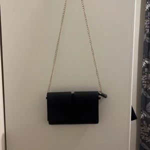 Svart axelväska med kedja - Snygg svart axelväska i syntetmaterial med en elegant kedjerem. Perfekt för både vardag och fest. Väskan har en stilren design med en enkel stängning framtill.