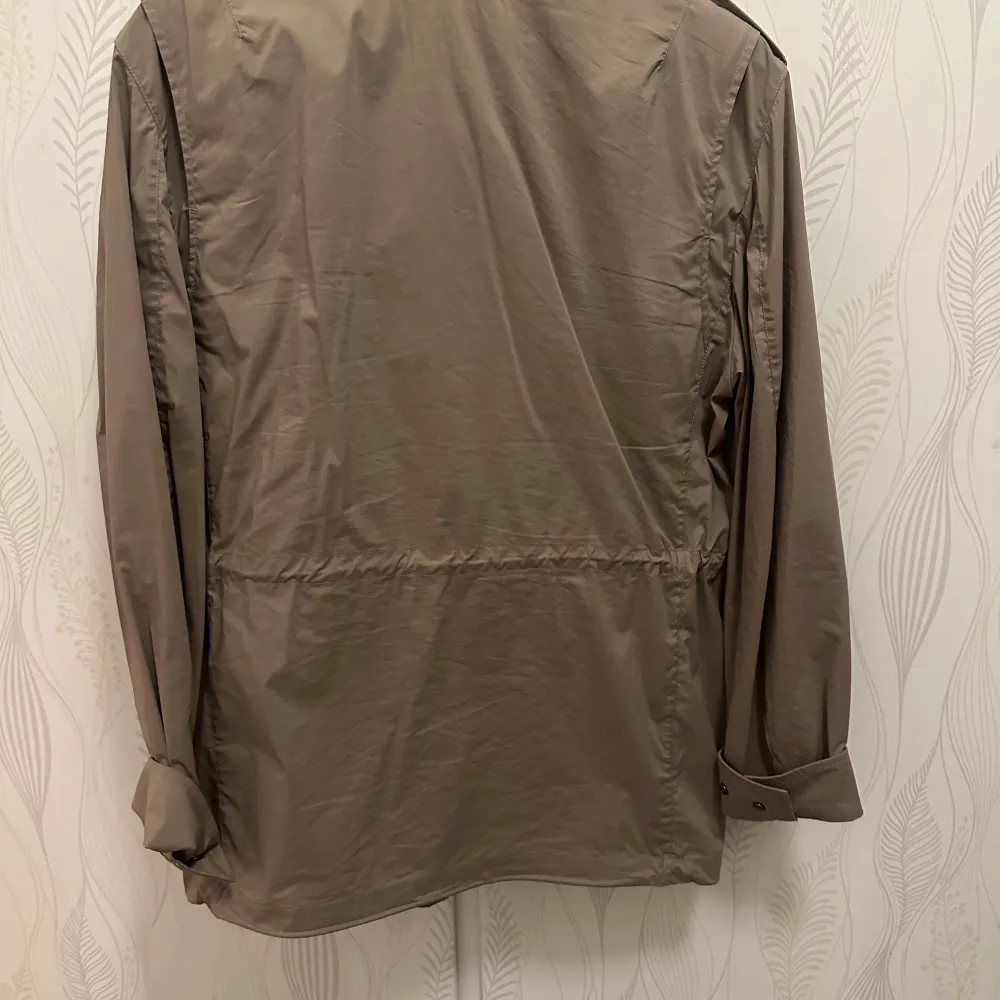 Säljer en beige jacka med dragkedja och knappar. Jackan har långa ärmar och flera praktiska fickor framtill. Perfekt för höst och vår. Inbyggd luva. Takit.