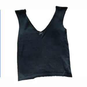 Svart ärmlös topp från Sisley - Snygg svart ärmlös topp från Sisley med en liten glittrig rosett-detalj vid halsringningen. Perfekt för både vardag och festliga tillfällen. Toppen har en djup v-ringning och är i ett mjukt material som känns skönt mot huden.