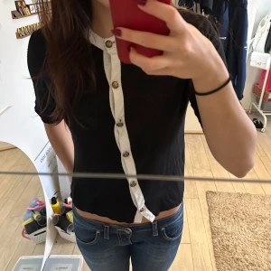 Svart kortärmad topp från Massimo Dutti - Säljer en elegant svart topp från Massimo Dutti i storlek XS. Den har korta ärmar och en vit knappslå med guldknappar som ger en snygg kontrast. Perfekt för både vardag och fest! 🖤
