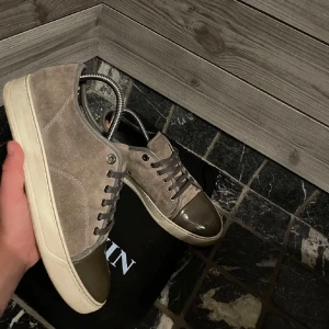 Lanvins sneaker -   Lanvin sneakers i mocka och läder Säljer ett par exklusiva Lanvin-sneakers i en stilren kombination av mocka och lackläder. Skorna har en tidlös design med:Gråbrun mocka, Lackad tåhätta i olivgrön. kommer med originalpåsen från Lanvin.
