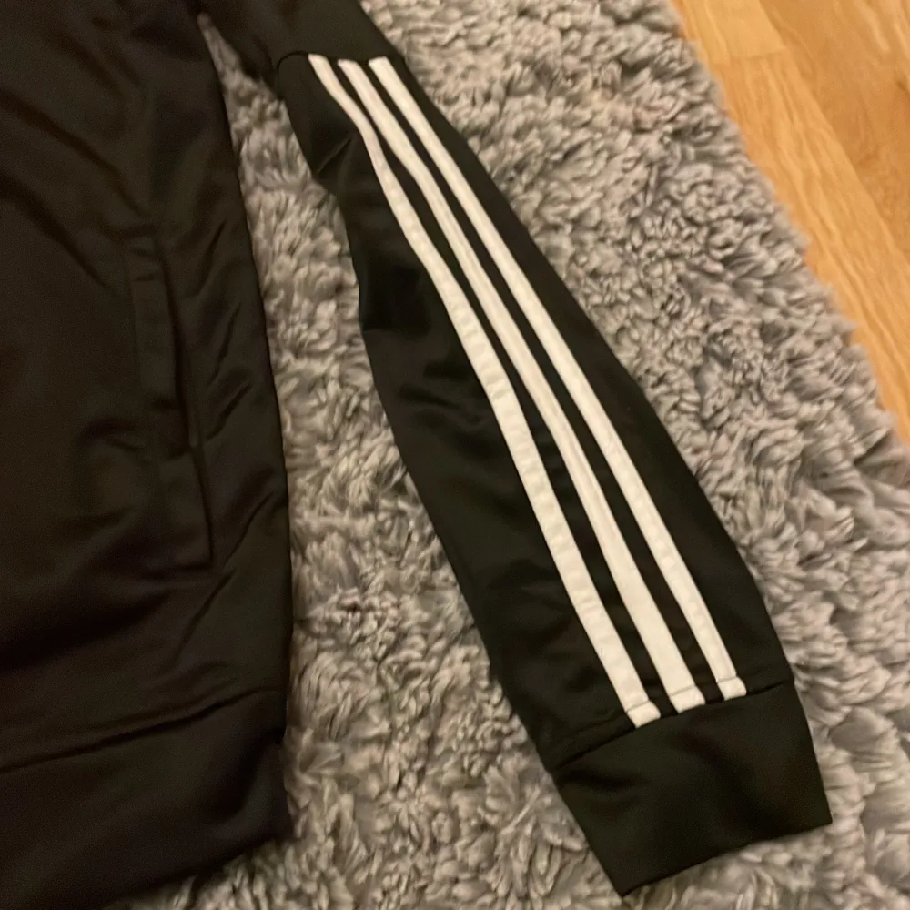 Snygg svart träningsjacka från Adidas med klassiska vita ränder längs ärmarna. Jackan har en dragkedja framtill och är tillverkad i ett syntetmaterial som ger en sportig look. Perfekt för träning eller en avslappnad stil.. Jackor.