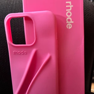 Trendigt mobilskal från Rhode! - Trendigt mörkrosa mobilskal i silikon med fäste för ditt favoritlipgloss. Passar dom flesta lipgloss och det behöver inte va från Rhode. Skalet passar IPhone 13/14/15 & PRO-modellerna till dom. Det är helt nytt och kommer med kartong. 