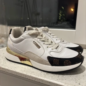 Vita sneakers från Guess med gulddetaljer - Snygga vita sneakers från Guess med gulddetaljer och svart mönster. Skorna har snörning och en stilren design som passar perfekt för en trendig look.