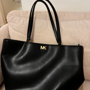 Svart axelväska från Michael Kors - Snygg och stilren svart axelväska från Michael Kors i skinn. Väskan har ett elegant MK-emblem i guld på framsidan och en praktisk dragkedja upptill. Insidan är rymlig med flera fack för enkel organisering. Perfekt för både vardag och fest!