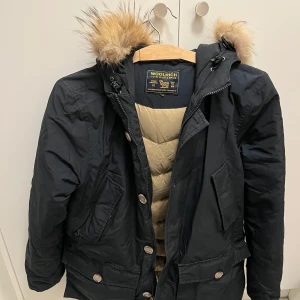 Marinblå parkajacka från Woolrich - Säljer en marinblå parkajacka från Woolrich med pälsdetaljer på huvan. Jackan har en dragkedja och knappar framtill samt flera praktiska fickor. Perfekt för kyliga dagar. Den är i storlek 14y men passar mig som har small i kläder och är 170 cm. Den är använd men är i gott skick (välbeverad).