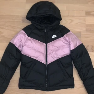 Pufferjacka från Nike - Snygg svart pufferjacka från Nike med rosa detaljer och huva. Jackan har en dragkedja framtill och Nike-logga på bröstet. Perfekt för vinter/höst. Har aldrig använt och i mycket bra skick 🤗Passar på mig perfekt som är 160 cm ifall någon undrar!!