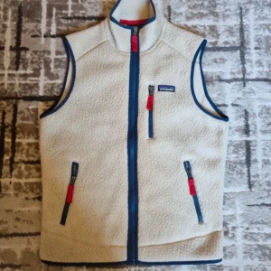 Beige fleeceväst från Patagonia - Säljer en beige fleeceväst från Patagonia med blå dragkedjor och röda detaljer. 