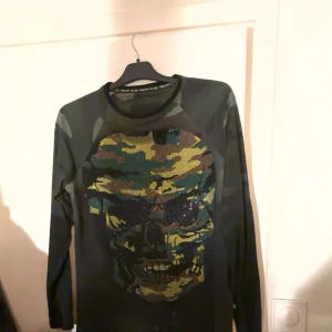 Camouflage tröja från Philipp Plein - Säljer en äkta sweatshirt tröja från Philipp Plein i camouflage-mönster. Den har en stor dödskalle i paljetter på framsidan och märkeslogga i rött på baksidan. Tröjan är i 100% bomull och perfekt för en edgy look. Passar bra till både vardag och fest!