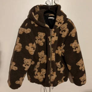 Brun teddyjacka med björnmönster - Mysig brun teddyjacka med gulligt björnmönster. Jackan har en dragkedja framtill och en huva för extra värme. Perfekt för kyliga dagar när du vill ha något både bekvämt och stiligt.
