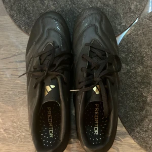 Svarta fotbollsskor Predator - Säljer ett par svarta Adidas Predator fotbollsskor med snörning. Skorna har en stilren design med diskreta detaljer och är perfekta för fotbollsplanen.