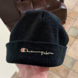 Svart mössa från Champion - 90-tal sotarmössa från Champion med broderad logga framtill. Perfekt för att hålla värmen under kyliga dagar. Enkel och stilren design som passar till det mesta.