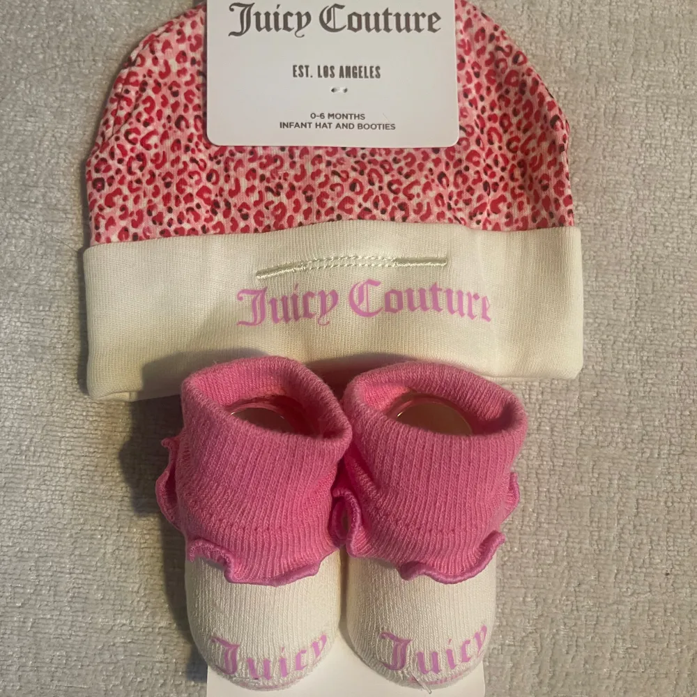 Supersött babyset från Juicy Couture med en mössa och matchande tossor. Mössan har ett rosa leopardmönster och vit kant med rosa text. Tossorna är rosa med volangdetaljer och vit bas med rosa text. Perfekt för att hålla din bebis varm och stilren!. Accessoarer.