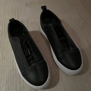Svarta sneakers i skinn - Snygga svarta sneakers i skinn med vit sula från Jim Rickey. De har en stilren design utan snörning, vilket gör dem enkla att ta på och av. Perfekta för en casual look och passar till det mesta i garderoben. Skorna är i nyskick (oanvända) och redo för nya äventyr!