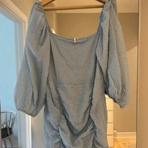 Blå miniklänning  - Blå tajt miniklänning med roucheade detaljer. Ballongärmar och fyrkantig urringning. 