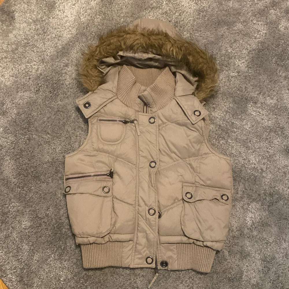 Säljer en superfin beige dunväst från Clockhouse i storlek S. Den har en mysig huva med pälsdetaljer och praktiska fickor med knappar och dragkedja. Perfekt för höst och vinter! 🧥✨. Takit.
