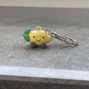 Gullig nyckelring med citronfigur - Söt nyckelring med en liten citronfigur som har ett glatt ansikte. Baserad på jellycat gosedjuren. Citronen är gul med gröna blad och små bruna fötter. Perfekt som en rolig accessoar till väskan eller nycklarna. Den är liten och lätt att bära med sig.