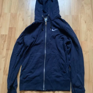 Mörkblå hoodie från Nike - Säljer en mörkblå hoodie från Nike i storlek S. Den har en dragkedja framtill och en klassisk Nike-logga på bröstet. Perfekt för höst och vinter, och den är superbekväm med en skön passform. Huvan har snören för justering. Perfekt för både träning och vardag!