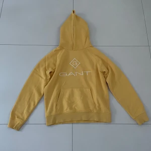 Gul hoodie från GANT - Säljer en snygg gul hoodie från GANT i storlek M. Den har en klassisk design med en stor ficka fram och GANT-loggan tryckt i vitt. Perfekt för höst och vår när du vill vara både bekväm och stilren. Huvan är mjuk och skön, och materialet känns riktigt bra mot huden.