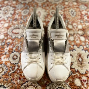 Valentino Garavani sneakers - Säljer ett par snygga Valentino Garavani sneakers i vitt med silverdetaljer. Skorna har en klassisk design med snörning och är i välanvänt skick. Perfekta för både vardag och festliga tillfällen!