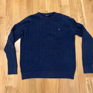 Mörkblå stickad tröja från GANT - Säljer en snygg mörkblå stickad tröja från GANT. Den har ett klassiskt kabelstickat mönster och är perfekt för både höst och vinter. Tröjan har långa ärmar och en rund halsringning. Perfekt för en stilren och bekväm look! (Nypris 900kr)