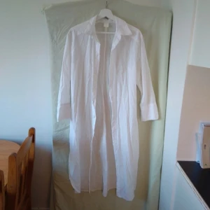 Vit lång skjortklänning - Säljer en stilren och enkel vit lång skjortklänning. Perfekt för en avslappnad sommarlook eller som ett lager över en annan outfit. Klänningen har långa ärmar och en klassisk krage. Passar bra till både vardag och festliga tillfällen.