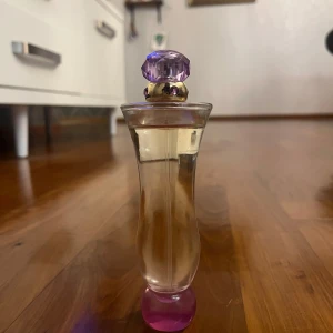 Versace Woman parfym 50ml - Elegant parfymflaska från Versace med en lila kristallformad kork och rosa bas. Flaskan har en smal, kurvig design som ger ett lyxigt intryck. Perfekt för den som vill ha en sofistikerad doftupplevelse. Förpackning medkommer ej.