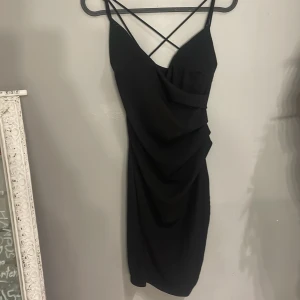 Svart klänning med korsade axelband - Snygg svart klänning med korsade axelband och draperad design. Perfekt för en kväll ute. Den har en tight passform och är tillverkad i ett mjukt material som ger en elegant look.Med en slits på sidan