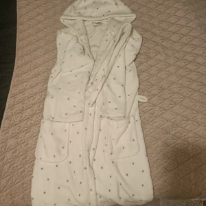 Vit fleece morgonrock med prickar - Mysig vit fleece morgonrock med små grå prickar och huva. Den har praktiska fickor och ett knytband i midjan för en bekväm passform. Perfekt för kalla morgnar eller mysiga kvällar hemma.