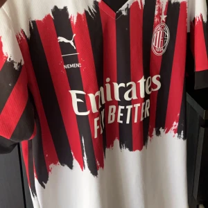 AC Milan fotbollströja från Puma - Snygg AC Milan fotbollströja från Puma med ett unikt röd-svart randigt mönster och vit bas. Tröjan har korta ärmar och klubbens emblem på bröstet.
