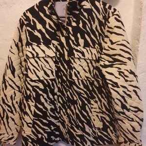 Zebramönstrad jacka med dragkedja - Säljer en cool vit och svart mönstrad jacka med dragkedja och fickor framtill. Jackan har långa ärmar och en avslappnad passform. Perfekt för att sticka ut i mängden!