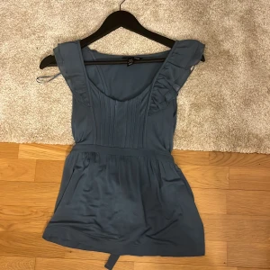 Blå ärmlös topp från H&M - Snygg blå ärmlös topp från H&M med volangdetaljer vid axlarna och knytband i midjan. Toppen har en elegant design med en mjuk och följsam passform. Perfekt för en stilren look.