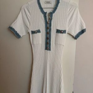 Vit ribbad klänning från Sandro - Snygg vit ribbad klänning från Sandro med korta ärmar och blå detaljer. Klänningen har knappar framtill och två fickor med dekorativa knappar. Perfekt för en stilren look.