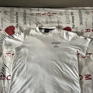 Vit t-shirt från Superdry - Säljer en stilren vit t-shirt från Superdry med svart text på bröstet och coola detaljer på ärmarna. Perfekt för en avslappnad look. T-shirten är kortärmad och gjord i mjukt material.
