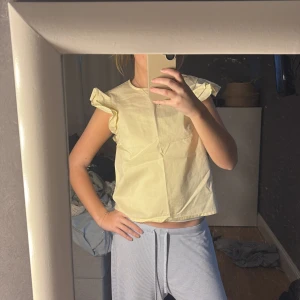 Gul blus med volangdetaljer - Söt gul blus med volangdetaljer vid ärmarna. Perfekt för en somrig look. Blusen har en enkel och stilren design som passar till många olika outfits.