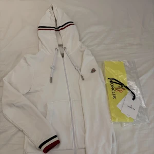 Moncler hoddie vit - Säljer en stilren vit hoodie från Moncler med dragkedja och huva. Den har snygga randiga detaljer i rött och svart på ärmsluten och huvan. Perfekt för en casual look.  Storlek 1 motsvarar S. Skriv för mer bilder 
