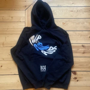 Svart hoodie från Marcus & Martinus - Säljer en svart hoodie från Marcus & Martinus med tryck i vitt och blått. Hoodien har en stor ficka framtill och en bekväm passform. Perfekt för en avslappnad stil.