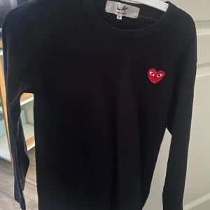 Svart långärmad tröja från Comme des Garçons - Säljer en svart långärmad tröja från Comme des Garçons med ett rött hjärta på bröstet. Tröjan är stilren och passar perfekt för en trendig look. Perfekt för dig som gillar minimalistisk design med en twist. Pris går att diskutera vid snabb affär.