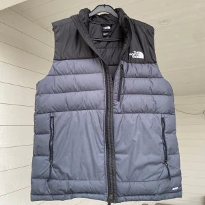 Svart dunväst från The North Face - Säljer en snygg svart dunväst från The North Face. Västen har en dragkedja framtill och två praktiska fickor med dragkedjor. Perfekt för kyligare dagar och ger en stilren look.