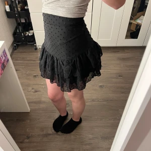 Svart kortkjol med volang - Snygg svart kortkjol med prickigt mönster och volangdetaljer. Perfekt för att ge en lekfull touch till din outfit. Kjolen har en tight passform i midjan och faller fint med sina lager av volanger. Stl xs