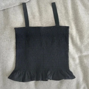 Svart smockad linne - Säljer en svart smockad linne med volangdetalj längst ner. Den har breda axelband och en elastisk passform som gör den bekväm att bära. Perfekt för varma dagar eller som ett lager under en jacka.