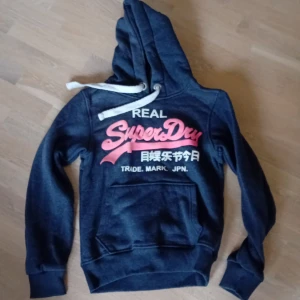 Mörkblå hoodie från Superdry - Säljer en mörkblå hoodie från Superdry med rosa och vit logga på framsidan. Den har en stor ficka framtill och justerbar huva med vita snören. Perfekt för en avslappnad stil.