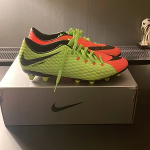 Nike Hypervenom fotbollsskor - Snygga Nike Hypervenom fotbollsskor i en iögonfallande kombination av neonorange och limegrön med svarta detaljer. Skorna har snörning och är designade för att ge bra grepp på planen. Perfekta för fotbollsspelare som vill sticka ut.